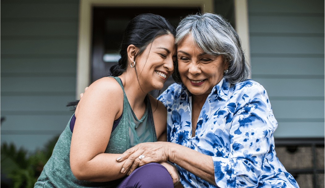 Photograph of an older woman and her adult daughter smiling and hugging each other on their front porch Photograph of an older woman and her adult daughter smiling and hugging each other on their front porch